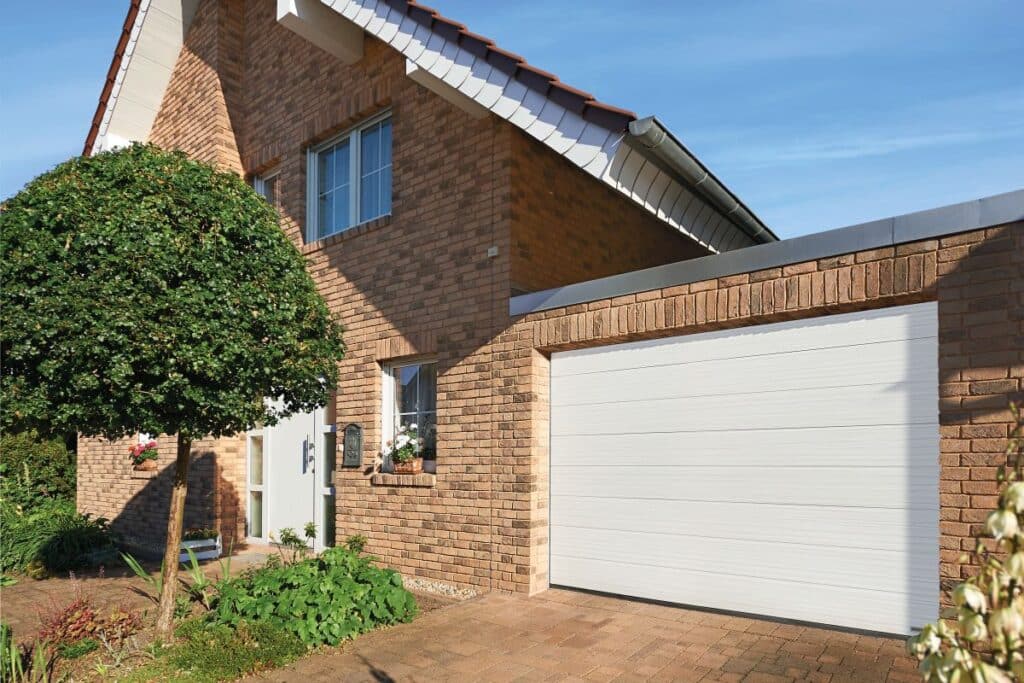 Backsteinhaus mit Garage, weißer Tür und einem gepflegten Baum. Gärten grenzen an die gepflasterte Auffahrt.