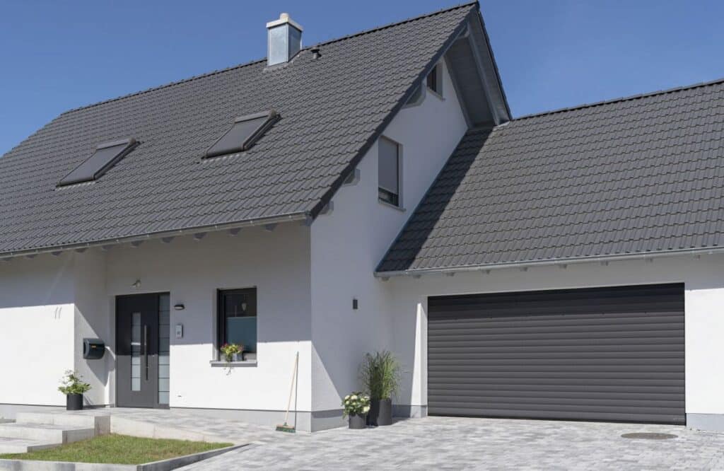 Modernes, zweistöckiges Haus mit grauem Dach, Garage und kleinem Garten davor. Blauer Himmel im Hintergrund.