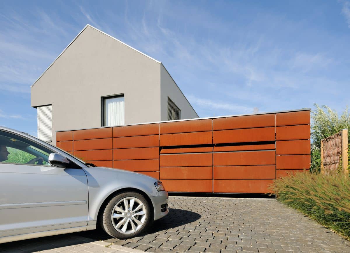 Modernes Hausäußeres mit rostfarbener Fassade, graues Auto parkt auf der gepflasterten Auffahrt unter einem klaren blauen Himmel.