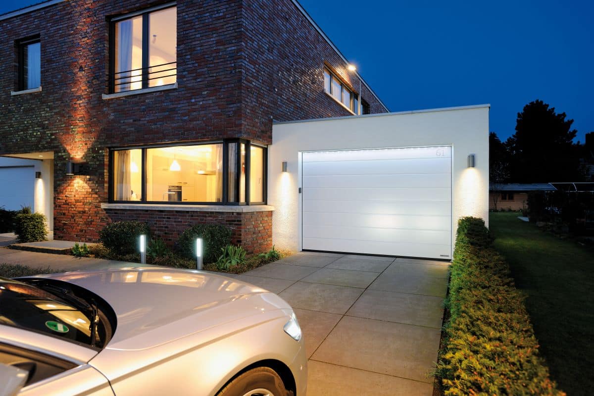 Modernes Haus mit beleuchtetem Garagentor in der Abenddämmerung, neben einem geparkten weißen Auto auf einer Betoneinfahrt.