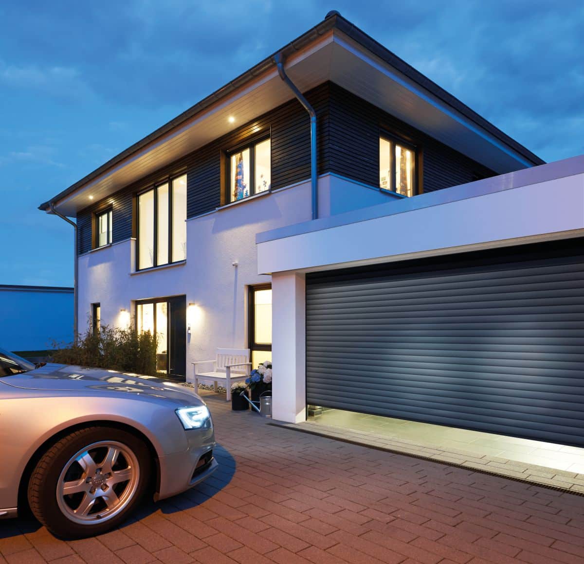 Modernes Haus mit großen Fenstern und einer in der Dämmerung beleuchteten Garage, in der Einfahrt parkt ein silbernes Auto.