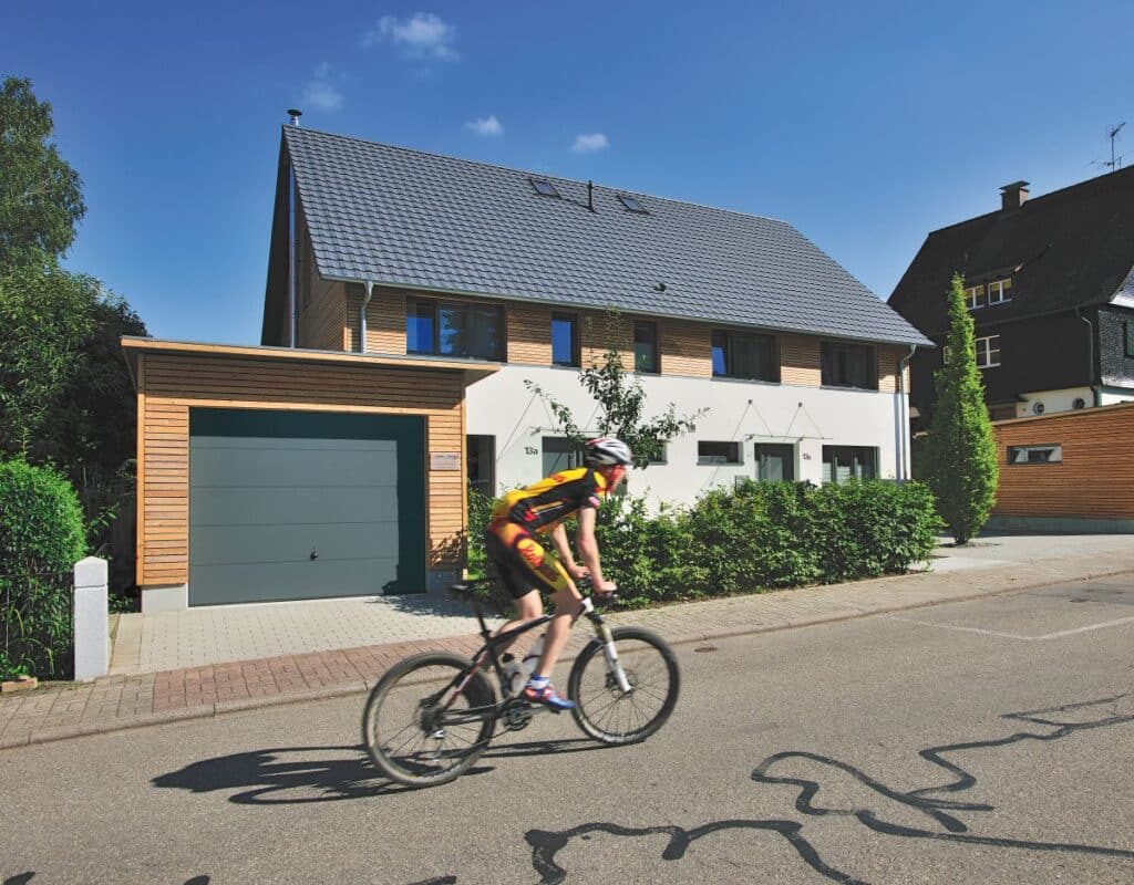 Radfahrer fährt unter einem klaren blauen Himmel an einem modernen Vorstadthaus mit Garagen-Schwingtor vorbei.