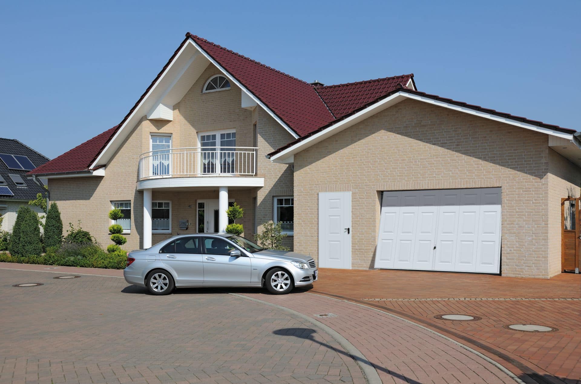 Ein modernes Haus mit rotem Dach, großen Fenstern und einem weißen Auto, das davor auf einer gepflasterten Auffahrt geparkt ist.