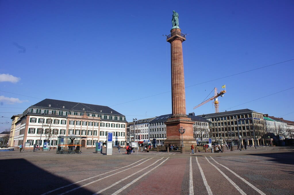 Eine hohe Säule mit einer Statue steht auf einem Stadtplatz, der von historischen und modernen Gebäuden umgeben ist.