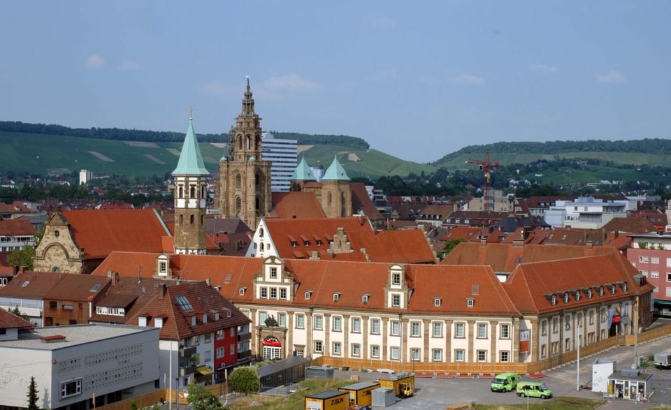 Gebäude mit roten Dächern und Kirchtürme in einer europäischen Stadt mit grünen Hügeln im Hintergrund unter einem blauen Himmel.