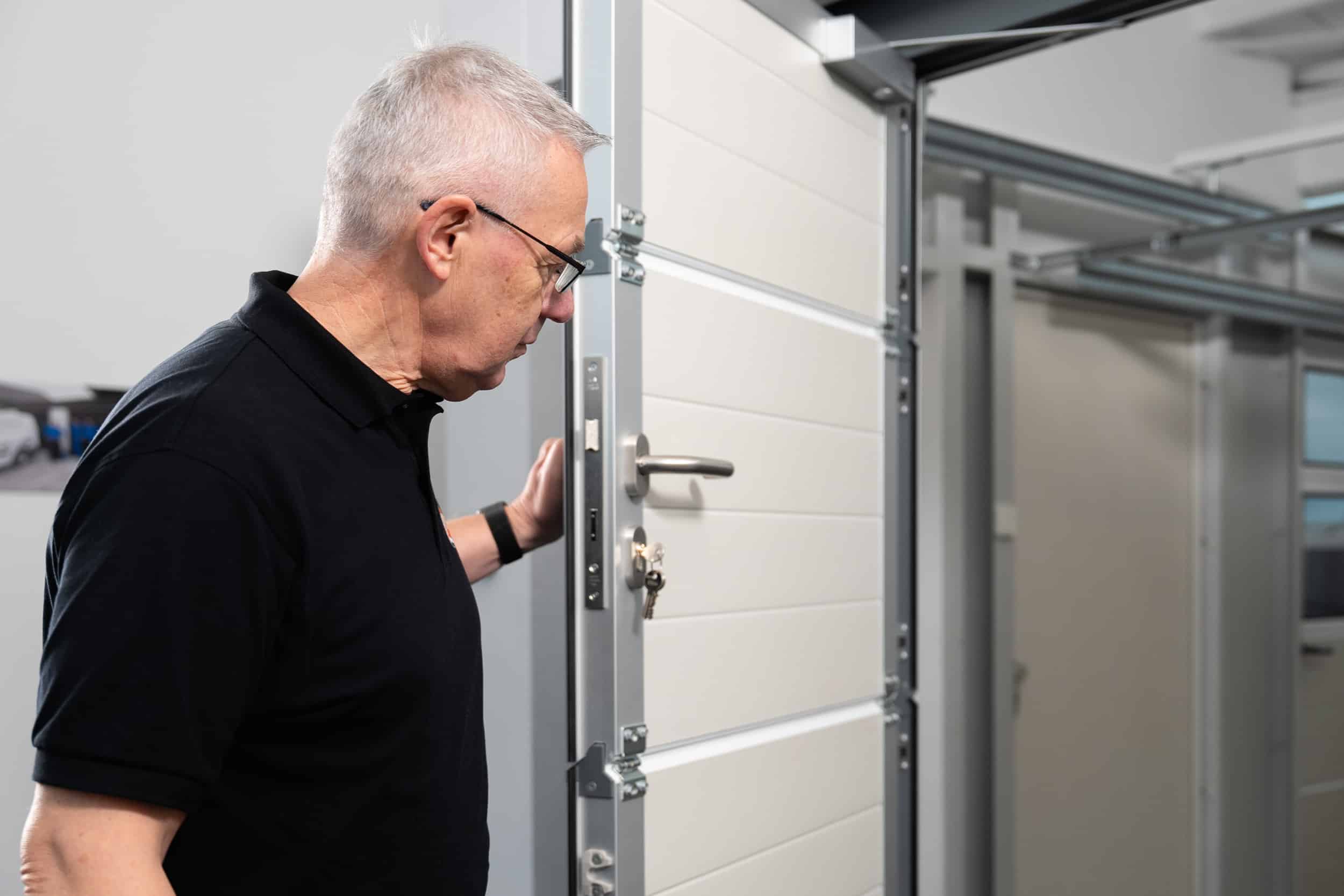 Älterer Mann mit Brille und schwarzem Hemd inspiziert eine weiße Tür in einem Innenraum.