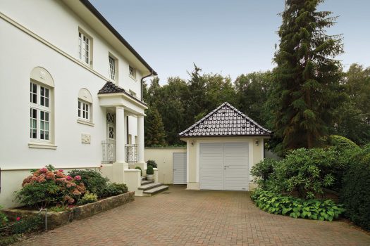 Weißes Haus mit Ziegeldach, angrenzender Garage, gepflasterter Auffahrt und üppigem Garten mit Sträuchern und Blumen davor.